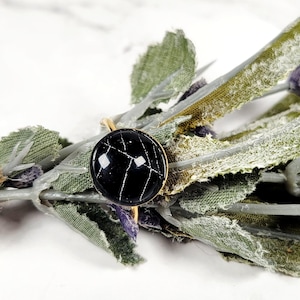 May include: A gold-colored ring with a round, black stone featuring white veining. The ring is set against a backdrop of green and purple dried flowers and leaves, with a natural twine accent.