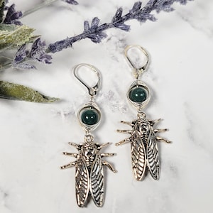 May include: Silver-tone earrings featuring a green bead and a detailed cicada charm. The earrings have a lever-back closure. The background includes sprigs of lavender and a marble-like surface.