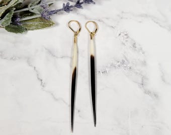 May include: A pair of long, slender earrings featuring natural porcupine quills that transition from a creamy white base to a dark brown, pointed tip. The quills are suspended from gold-toned metal lever-back ear wires, creating a dangling design.