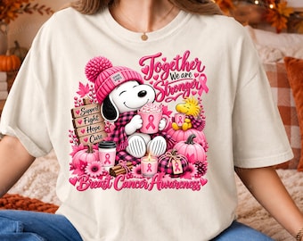Könnte beinhalten: Ein cremefarbenes T-Shirt mit einer Grafik eines Zeichentrickhundes in rosa Mütze und Schal, der eine rosa Tasse hält. Das Design zeigt rosa Kürbisse, ein Holzschild mit der Aufschrift Support, Fight, Hope, Cure und den Text Together We Are Stronger Breast Cancer Awareness.