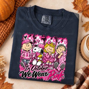 K&ouml;nnte beinhalten: Ein dunkelblaues T-Shirt mit einer Grafik von vier Peanuts-Figuren in rosa Kleidung und Accessoires. Das Design enth&auml;lt rosa Schleifen zur Brustkrebsaufkl&auml;rung und den Text "In October We Wear Pink" in einer fetten, stilisierten Schrift.