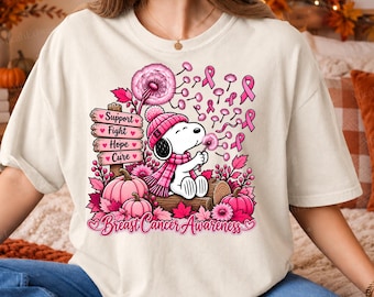 Könnte beinhalten: Ein cremefarbenes T-Shirt mit einer Grafik eines Zeichentrickhundes, der eine rosa Strickmütze und einen Schal trägt und auf einem Baumstamm sitzt. Das Design zeigt rosa Kürbisse, Blätter, Pusteblumen, Schleifen und Schilder mit der Aufschrift Support, Fight, Hope, Cure, Breast Cancer Awareness.