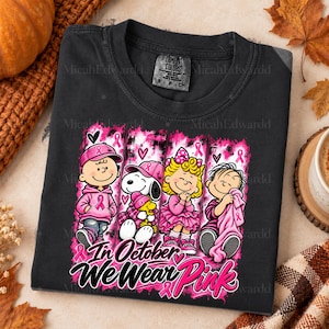K&ouml;nnte beinhalten: Ein schwarzes T-Shirt mit einer Grafik von vier Zeichentrickfiguren, die rosa Kleidung und Accessoires sowie rosa Schleifen tragen. Der Text unter den Figuren lautet In October We Wear Pink. Das Shirt hat ein Comfort Colors Etikett.