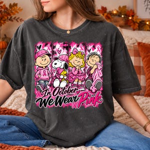 K&ouml;nnte beinhalten: Ein anthrazitfarbenes T-Shirt mit einem Grafikdruck von vier Zeichentrickfiguren in rosa Kleidung und Accessoires. Das Design enth&auml;lt rosa Schleifen und den Text "In October We Wear Pink" in einer fetten, schwarzen und rosa Schriftart.