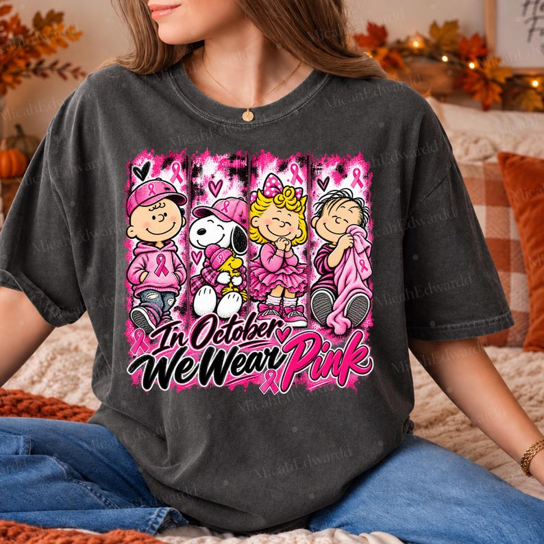 K&ouml;nnte beinhalten: Ein anthrazitfarbenes T-Shirt mit einem Grafikdruck von vier Zeichentrickfiguren in rosa Kleidung und Accessoires. Das Design enth&auml;lt rosa Schleifen und den Text "In October We Wear Pink" in einer fetten, schwarzen und rosa Schriftart.