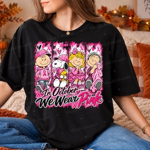 K&ouml;nnte beinhalten: Ein schwarzes T-Shirt mit einem Grafikdruck von vier Zeichentrickfiguren in rosa Kleidung und Accessoires. Das Design enth&auml;lt rosa Schleifen und den Text In October We Wear Pink. Die Figuren sind in vertikalen Feldern angeordnet.