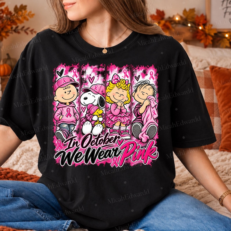 K&ouml;nnte beinhalten: Ein schwarzes T-Shirt mit einem Grafikdruck von vier Zeichentrickfiguren in rosa Kleidung und Accessoires. Das Design enth&auml;lt rosa Schleifen und den Text In October We Wear Pink. Die Figuren sind in vertikalen Feldern angeordnet.