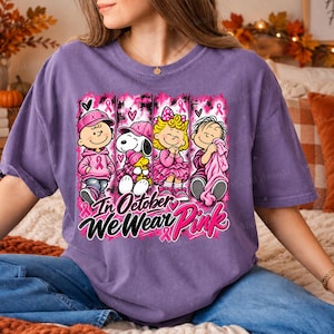 K&ouml;nnte beinhalten: Ein violettes Kurzarm-T-Shirt mit einer Grafik von vier Zeichentrickfiguren in rosa Kleidung und Accessoires. Das Design enth&auml;lt rosa Schleifen und den Text In October We Wear Pink in schwarzer und rosa Schrift und Blockbuchstaben.