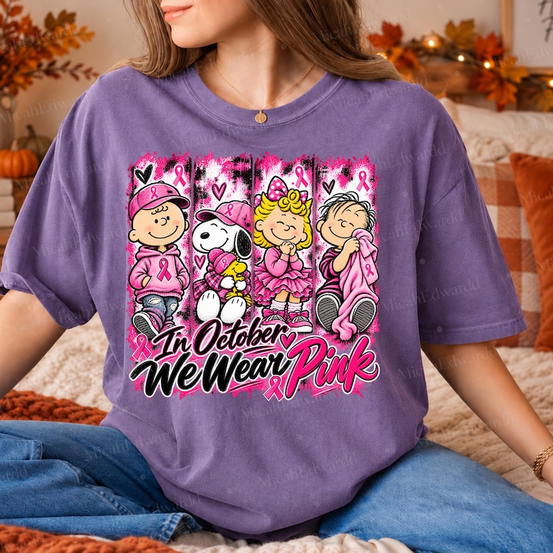 K&ouml;nnte beinhalten: Ein violettes Kurzarm-T-Shirt mit einer Grafik von vier Zeichentrickfiguren in rosa Kleidung und Accessoires. Das Design enth&auml;lt rosa Schleifen und den Text In October We Wear Pink in schwarzer und rosa Schrift und Blockbuchstaben.