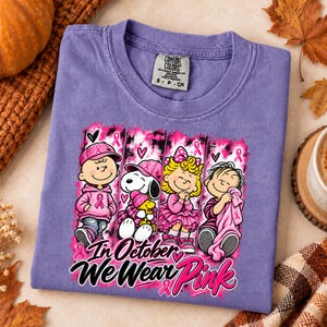 K&ouml;nnte beinhalten: Ein violettes Kurzarm-T-Shirt mit einer Grafik von vier Zeichentrickfiguren in rosa Kleidung und Accessoires. Der Text lautet In October We Wear Pink. Das Design enth&auml;lt rosa Schleifen, Herzen und einen Hintergrund mit Pinselstrich-Optik.