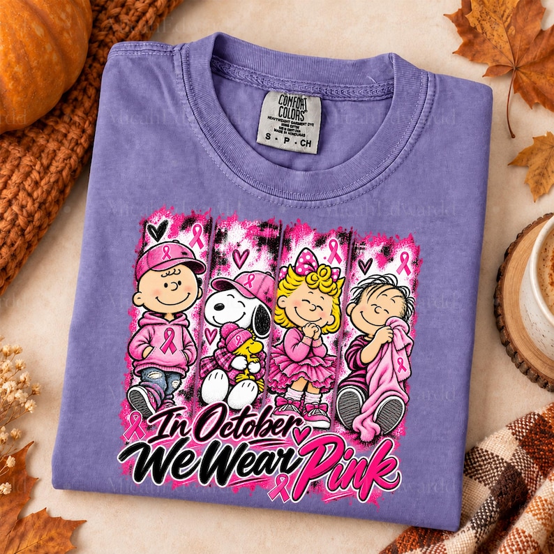 K&ouml;nnte beinhalten: Ein violettes Kurzarm-T-Shirt mit einer Grafik von vier Zeichentrickfiguren in rosa Kleidung und Accessoires. Der Text lautet In October We Wear Pink. Das Design enth&auml;lt rosa Schleifen, Herzen und einen Hintergrund mit Pinselstrich-Optik.