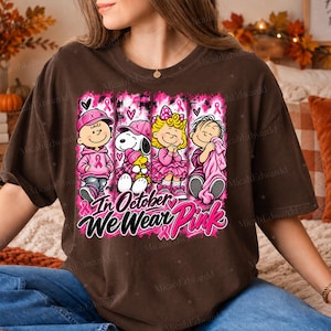 K&ouml;nnte beinhalten: Ein dunkelbraunes T-Shirt mit einer Grafik von vier Zeichentrickfiguren, die rosa Kleidung und Accessoires tragen. Das Design enth&auml;lt rosa Schleifen und den Text "In October We Wear Pink" in einer stilisierten schwarz-rosa Schrift.