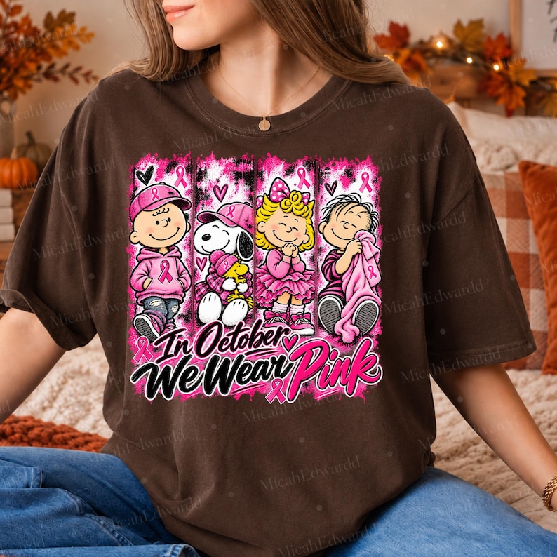 K&ouml;nnte beinhalten: Ein dunkelbraunes T-Shirt mit einer Grafik von vier Zeichentrickfiguren, die rosa Kleidung und Accessoires tragen. Das Design enth&auml;lt rosa Schleifen und den Text "In October We Wear Pink" in einer stilisierten schwarz-rosa Schrift.