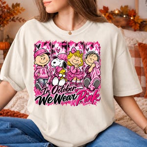 K&ouml;nnte beinhalten: Ein cremefarbenes T-Shirt mit einer Grafik von vier Zeichentrickfiguren in rosa Outfits und Accessoires. Das Design enth&auml;lt rosa Schleifen und den Text In October We Wear Pink. Die Figuren sind in vertikalen Feldern mit rosa Akzenten angeordnet.