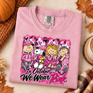 K&ouml;nnte beinhalten: Ein hellrosa T-Shirt mit einer Grafik von vier Zeichentrickfiguren in rosa Kleidung und Accessoires. Das Design enth&auml;lt rosa Schleifen und den Text In October We Wear Pink. Am Halsausschnitt befindet sich ein Comfort Colors Markenetikett.