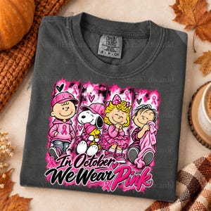 K&ouml;nnte beinhalten: Ein anthrazitfarbenes T-Shirt mit einer Grafik von vier Zeichentrickfiguren in rosa Kleidung und Accessoires. Das Design enth&auml;lt rosa Schleifen und den Text "In October We Wear Pink" in einer fetten, stilisierten Schriftart.