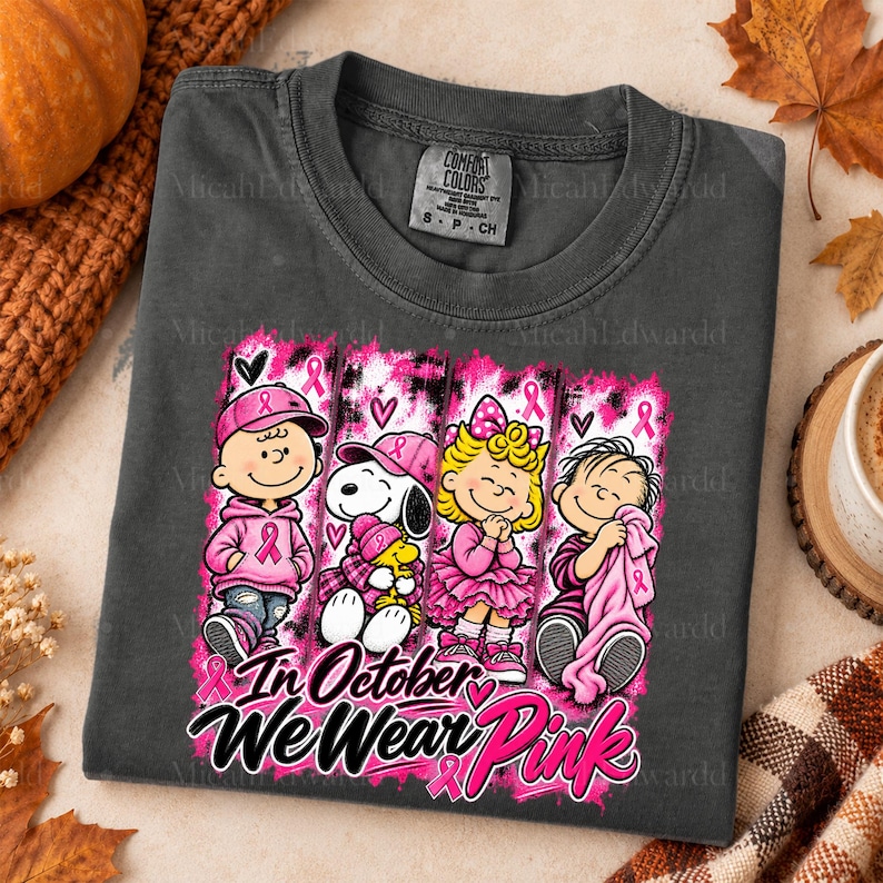 K&ouml;nnte beinhalten: Ein anthrazitfarbenes T-Shirt mit einer Grafik von vier Zeichentrickfiguren in rosa Kleidung und Accessoires. Das Design enth&auml;lt rosa Schleifen und den Text "In October We Wear Pink" in einer fetten, stilisierten Schriftart.