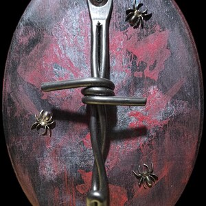 May include: Oval wooden wall art with a dark red and black painted background. A twisted metal piece is affixed to the wood, with three small metal spider accents. The metal piece has a fork-like end.