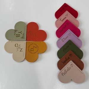 May include: A collection of heart-shaped and four-leaf clover-shaped leather bookmarks in various colors. The bookmarks feature personalized initials and names such as "Diana" and "Marisol", with decorative stitching.