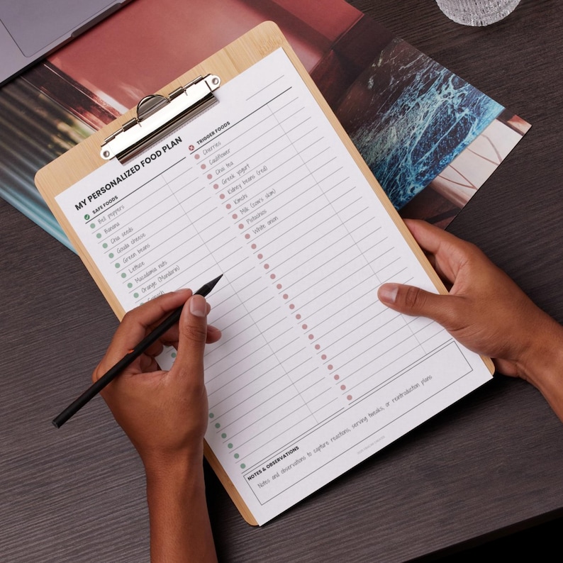 May include: A wooden clipboard with a printed "MY PERSONALIZED FOOD PLAN" form. The form lists safe and trigger foods. A person is holding the clipboard and writing with a black pen. The clipboard has a silver metal clip.