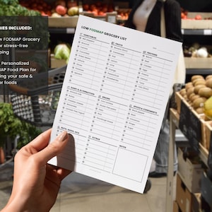 May include: A hand holds a white Low FODMAP Grocery List in a grocery store. The list includes sections for vegetables, fruits, proteins, dairy, grains, spices, and extras. The text "FREEBIES INCLUDED" is visible, along with text about a food plan.