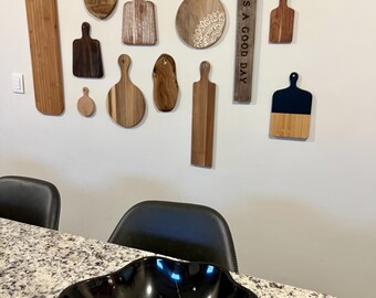 May include: A black marble bowl with a wavy rim sits on a granite countertop. Above, a collection of wooden cutting boards of various shapes and sizes adorns the wall. A wooden sign reads "TODAY IS A GOOD DAY."