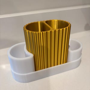 May include: A gold-colored, ribbed toothbrush holder with two compartments sits on a white tray with two additional side compartments. The holder is designed to organize bathroom essentials. The design is modern and functional.