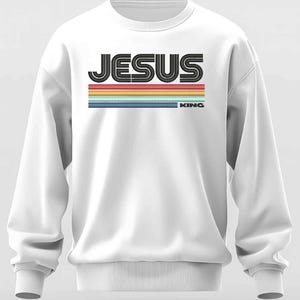 May include: White sweatshirt with the word "JESUS" in a black, retro font, above horizontal stripes in red, yellow, blue, and purple. The word "KING" is below the stripes. The sweatshirt has long sleeves and a ribbed hem and cuffs.