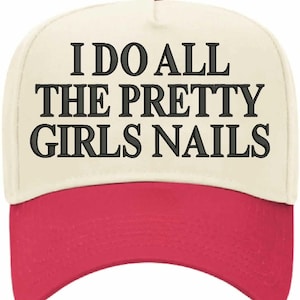 May include: A baseball cap with a cream-coloured crown and a red brim. The cap features the embroidered text "I DO ALL THE PRETTY GIRLS NAILS" in black block letters. The cap has a red button on top.