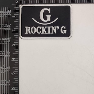 Könnte beinhalten: Schwarzer rechteckiger Aufnäher mit weiß gesticktem Text und einem stilisierten "G" über den Worten "ROCKIN' G". Der Aufnäher ist etwa 7,6 cm breit und 5 cm hoch.