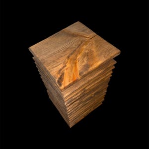 May include: A stack of wooden planks, forming a square column. The wood has a natural, warm brown tone with visible grain and knots. The edges of the planks are slightly uneven, adding to the rustic aesthetic. The background is solid black.