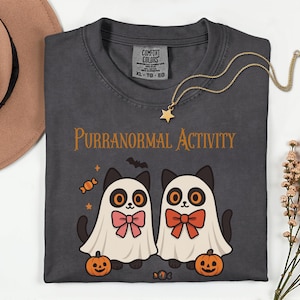 Puede incluir: Camiseta gris oscuro con la frase "Purranormal Activity" y un gráfico de dos gatos de dibujos animados disfrazados de fantasmas. Los gatos tienen ojos naranjas, pajaritas rosas y naranjas e ilustraciones de Halloween.