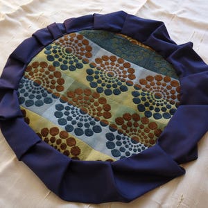 May include: A round, navy blue cushion with a ruffled edge. The cushion has a patterned fabric with brown, blue, and yellow swirls.