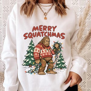 May include: A white long-sleeve sweatshirt featuring a graphic of a brown Bigfoot wearing a red and white patterned sweater. The creature is tangled in colorful Christmas lights between two pine trees. The text MERRY SQUATCHMAS is printed in red.