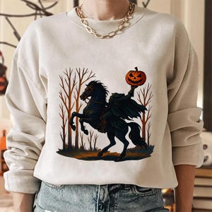 Może przedstawiać: Beżowa bluza z motywem Halloween. Projekt przedstawia bezgłowego jeźdźca na czarnym koniu w lesie, trzymającego rzeźbioną pomarańczową dynię. Grafika ma mroczną, upiorną estetykę, idealną na jesień.