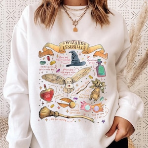 May include: White sweatshirt with a colorful graphic of wizarding essentials. The design includes a wizard hat, owl, potion bottles, broom, and text that reads "Wizard Essentials". The sweatshirt has a crew neck and long sleeves.