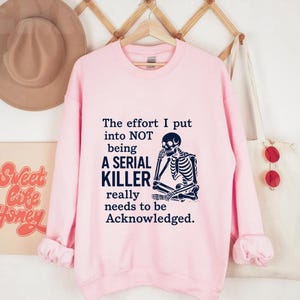 May include: A pale pink sweatshirt featuring the text "The effort I put into NOT being A SERIAL KILLER really needs to be Acknowledged." A black skeleton graphic is on the right side of the text.