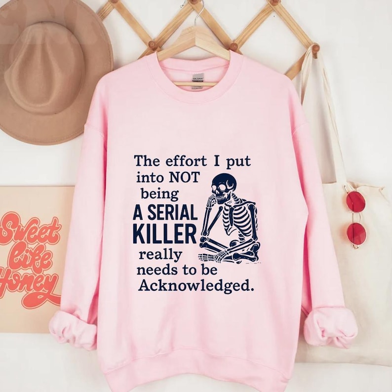 May include: A pale pink sweatshirt featuring the text "The effort I put into NOT being A SERIAL KILLER really needs to be Acknowledged." A black skeleton graphic is on the right side of the text.