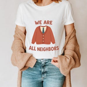 Op de afbeelding: Wit T-shirt met de tekst "WE ARE ALL NEIGHBORS" in bruin. Het shirt heeft een cartoon illustratie van een roestkleurig vest met een groene stropdas. De persoon draagt een blauwe spijkerbroek en een beige vest.