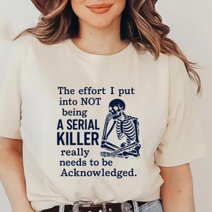 May include: A cream-coloured t-shirt featuring the text "The effort I put into NOT being A SERIAL KILLER really needs to be Acknowledged." A blue skeleton graphic is also on the shirt.