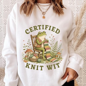 May include: White sweatshirt with a whimsical graphic of a frog knitting a colorful scarf. The frog is seated on a wooden chair, surrounded by mushrooms and foliage. The text "CERTIFIED KNIT WIT" is arched above and below the frog.