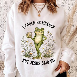 May include: A white long-sleeve sweatshirt featuring a graphic of a green frog holding a coffee mug, surrounded by wildflowers and mushrooms. The text "I COULD BE MEANER BUT JESUS SAID NO" is printed in black capital letters above and below the frog.