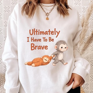 May include: White sweatshirt with the text "Ultimately I Have To Be Brave" in orange. The graphic shows two cartoon monkeys, one walking and pulling the other. The walking monkey is gray, and the other is orange.