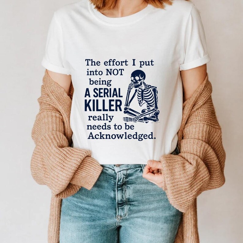 May include: White t-shirt with the text "The effort I put into NOT being A SERIAL KILLER really needs to be Acknowledged." and a blue skeleton graphic. The shirt is worn with blue jeans and a beige cardigan.