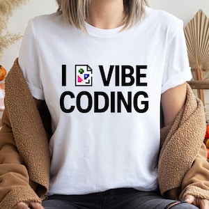 Peut inclure: T-shirt blanc avec l'inscription noire "I 💻 VIBE CODING". L'icône "💻" est un design pixélisé avec des carrés verts, bleus et roses. Le t-shirt est porté avec une veste marron.