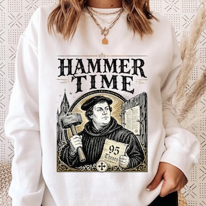 May include: White sweatshirt featuring a graphic of a man holding a hammer and a paper with the text "95 Theses." The design includes the words "Hammer Time" in a vintage font, with a church and wooden door in the background.