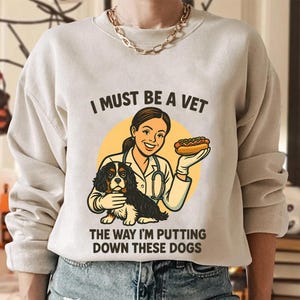 Może przedstawiać: Beżowa bluza z napisem "I MUST BE A VET THE WAY I'M PUTTING DOWN THESE DOGS". Grafika przedstawia weterynarza trzymającego hot doga i Cavalier King Charles Spaniel. Weterynarz ma na sobie biały fartuch i stetoskop.