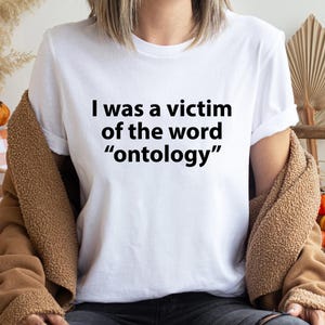 May include: A white short-sleeved t-shirt featuring the black printed text I was a victim of the word ontology. The shirt has a crew neckline and a standard fit, presented as a plain garment without any additional patterns or graphic designs.