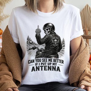 以下が含まれることがあります： バイクに乗った骸骨が中指を立てている白いTシャツ。バイカーは「RIDE FREE LIVE FREE」の文字が入った革ジャンを着ています。シャツには「CAN YOU SEE ME BETTER IF I PUT UP MY ANTENNA」の文字が入っています。
