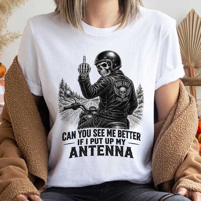 以下が含まれることがあります： バイクに乗った骸骨が中指を立てている白いTシャツ。バイカーは「RIDE FREE LIVE FREE」の文字が入った革ジャンを着ています。シャツには「CAN YOU SEE ME BETTER IF I PUT UP MY ANTENNA」の文字が入っています。